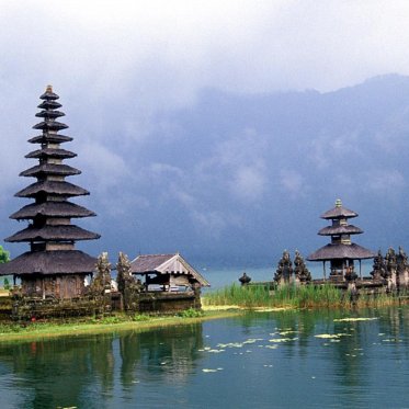 Photo of a small outcropping in water with a large and a small temple in Bali with mist and mountains in the backdrop.