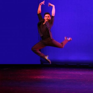A man dressed in black photographed in the air, posing for a dance while against a dark blue backdrop.
