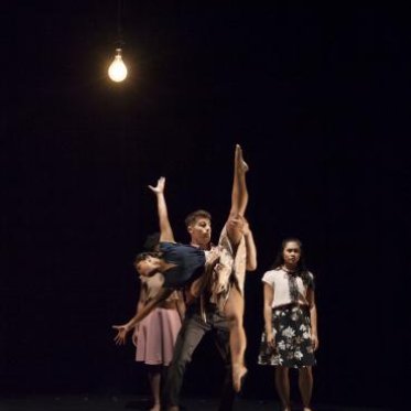A woman a flexible dance move, assisted by a male dancer. Two people stand rigidly against a black backdrop. A single lightbulb on top of the frame illuminates them.