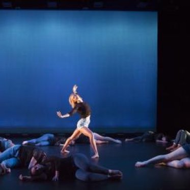 A blonde woman dances on the stage among still bodies on the stage floor