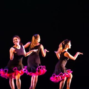 Three woman dressed identically, black clothing with pink at the tails of her skirt, with their arms extended during a dance performance.