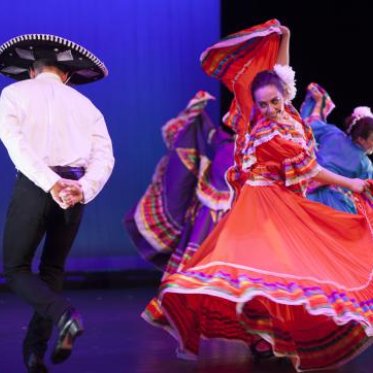 On the right, a woman dressed in traditional folklorico dress mid-dance. On the left, the back of a male dancer wearing a sombrero