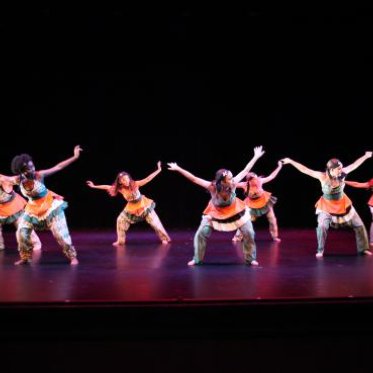 Seven woman wearing vibrant garments dance on the stage. 