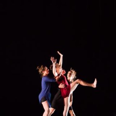 Two women surround a woman in red, standing on one leg and raising her hands up towards the air during a dance performance