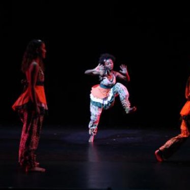 A woman is dancing while being lit by a bright spotlight, while the other dancers are in the dark, lit by an overhead red light that fades them into the black backdrop
