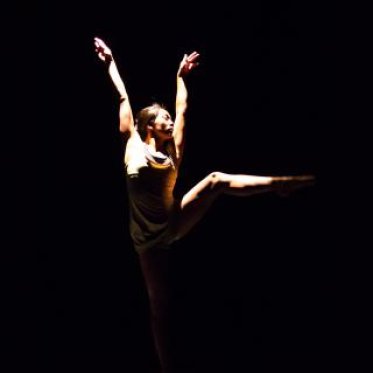 A woman in white, lit by a single spotlight from above, is performing a dance