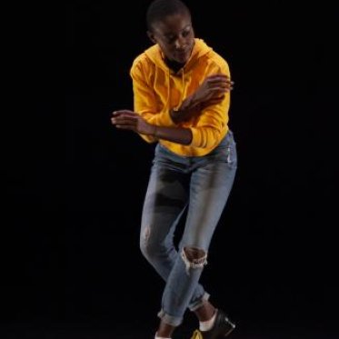 A woman in a yellow hoodie and a pair of jeans in the middle of a dance pose, her arms halfway crossed and bending her knees slightly. She is standing in front of a black background.