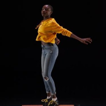 A woman in a yellow hoodie and a pair a jeans standing a wooden cutout on the floor. Her arms are extending backwards. She is set against a black background. 