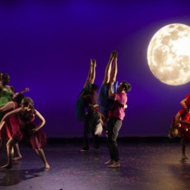 A group of dancers wearing bright colors dance against a blue background, with a superimposed and detailed picture of the moon to the right of the frame. Some dancers are by themselves to the far left of the frame. To the middle and the right, some dancers are holding other dances upside down. 