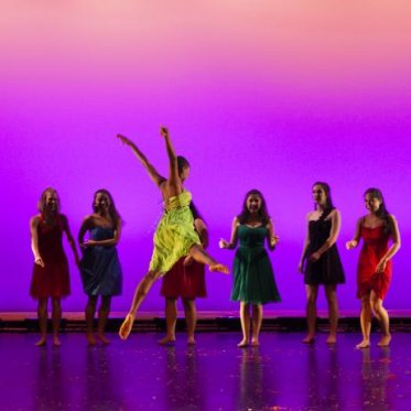 A woman in a yellow dress jumps across the stage during a dance performance while other woman in colorful dresses watch amazed