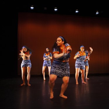 Five women in similar garments of blue and orange with a distinct pattern performing a dance. 